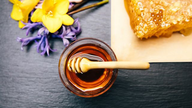 Healthy, natural, organic and sweet honey in glass jar or bowl with honey dipper, flowers and honeycomb on black background.