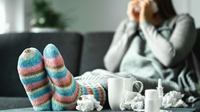 Sick woman with flu, cold, fever and cough sitting on couch at home. Ill person blowing nose and sneezing with tissue and handkerchief. Woolen socks and medicine. Infection in winter. Resting on sofa.